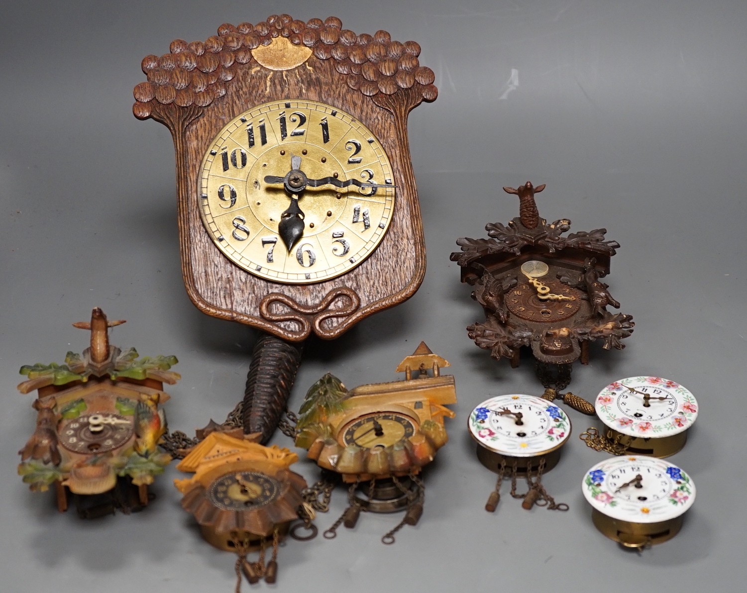 An early 20th century German Jugendstil oak wall timepiece, four miniature Black Forest timepieces and three floral enamelled examples, largest 23cms high